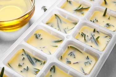 Ice cube tray with herbs frozen in oil and fresh rosemary on grey table, closeup