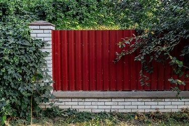 Fence made of panels of red corrugated sheet metal on a retaining wall with trees behind it.