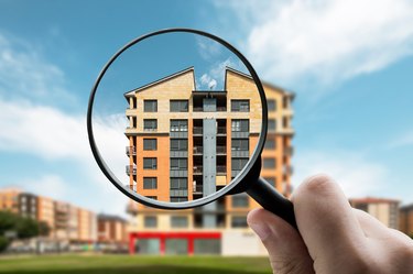 A hand holding up a magnifying glass that is zooming in on an apartment building in front of a blue sky with clouds.