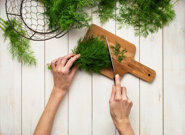 Top view of a woman's hand, fresh dill on a cutting board, knife and bunches of dill on a white wooden table. Fresh greens, seasoning, the process of cooking, a healthy lifestyle.