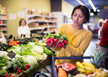 Woman wearing a yellow sweater picks ripe radishes off shelf in grocery store.