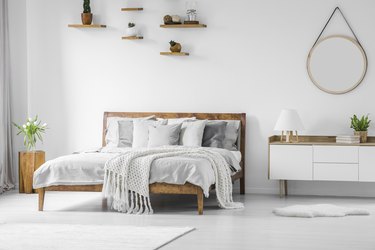 Comfortable big wooden frame bed with bed linens, pillows and blanket, nightstand beside and round mirror hanging on a white wall in a bright bedroom interior. Real photo.