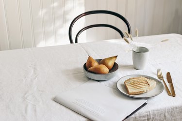 Breakfast with pears, toast, folded newspaper, and dried plants in mug on white linen table cloth.