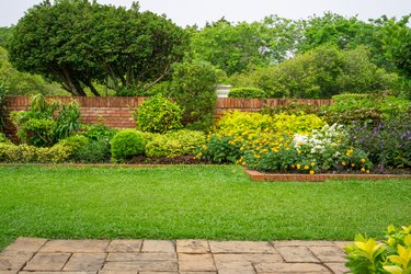 Backyard English cottage-style garden, colorful flowering plants and green grass lawn, brown pavers, and brick wall.