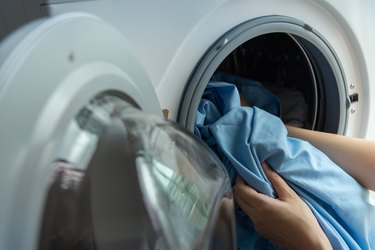 Woman’s hand putting dirty blue bed sheets into white washing machine.