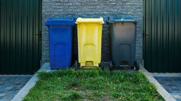 Blue, yellow, and gray trash bins standing in a row in backyard.