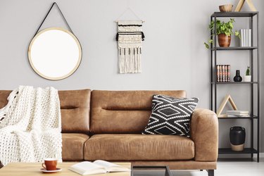 Real photo of handmade macrame and mockup poster hanging on the wall in bright sitting room interior with leather sofa and metal rack with decor and books.