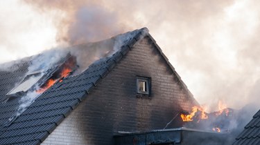 Fragment of a sooty white brick home that is on fire with flames and smoke cming out