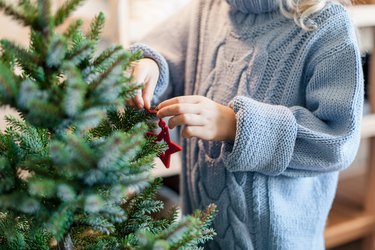 Little child girl is decorating Christmas tree with red ornaments. Kid is wearing in cotton knitted blue sweater.