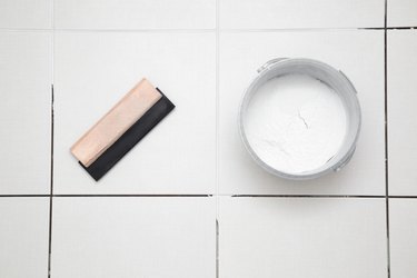 Rubber trowel and container with powder of grouting paste for ceramic tile seams on floor. Closeup. Top down view.