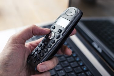 Hand Holding a Black Cordless Telephone with Laptop at Background