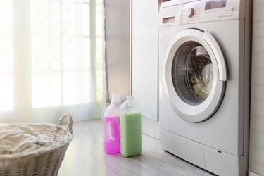 A front-loading washing machine in a laundry room next to green and pink laundry detergent and a laundry basket