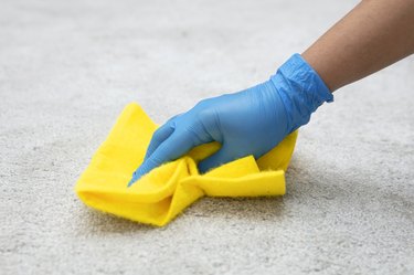 Person in blue gloves blotting a spill with a bright yellow cleaning cloth on white carpet.