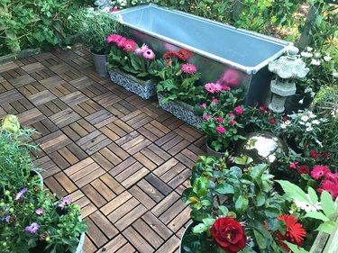Image of garden treehouse terrace platform balcony in summer with zinc metal raised pond water feature of galvanised cattle trough ready for water, plants, fountain, red miniature roses, pink gerbera flowers, wooden decking tiles, solar lights lighting