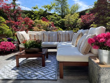 Image of outdoor lounging area on garden decking with outdoor rug, grooved, whitewashed wooden deck, hardwood seating with cushions, bonsai trees, Japanese maples, landscaped oriental design garden, focus on foreground.