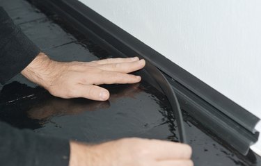 A man is installing a black PVC skirting board along a white wallpapered wall and hardwood painted floor to conceal electric cables behind a plastic moulding..