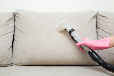 Person wearing protectives gloves using spray nozzle of professional vacuum cleaner and washing light beige back pillow of sofa at home. Extraction method. Commercial cleaning service. Closeup.