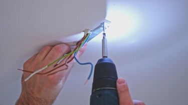 Caucasian Male Handyman Using Electric Screwdriver to Install Ceiling Lamp Mounting Plate Close to Power Cables