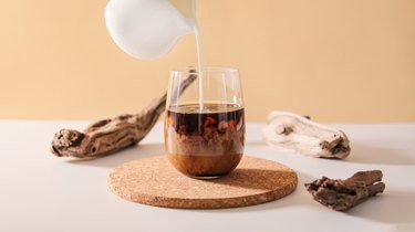 Milk cream pours into a transparent glass of black coffee on a minimalistic beige natural wooden background.