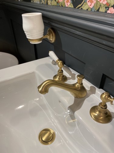 Close-up image of bronze mixer tap and toothbrush holder over a rectangular, white ceramic sink, wood panelling painted black and floral wallpaper in a luxury bathroom