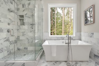Luxury master bathroom with elegantly tiled shower in addition to a soaking bathtub installed below a large window with view of trees.