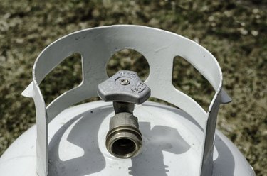 Angled overhead close-up of the top of a white propane tank and brass valve with grass in the background