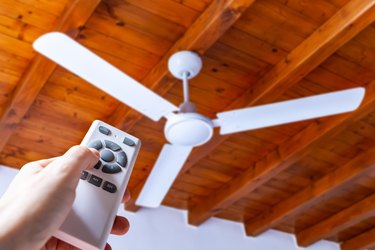 Close up shot of a hand using a remote control to operate a ceiling fan mounted in a house on a wooden ceiling.