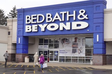 A Bed Bath & Beyond store with a royal blue entrance and white logo, with two shoppers entering the store from the parking lot.