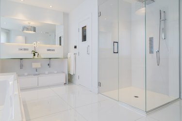 Detail of white rectangular bathtub, wall mirror, glass shower stall and sauna room door in en suite with ceramic tile flooring, upstairs inside luxury residential home