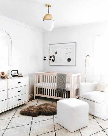 All-white gender neutral nursery with white armchair, white dresser, blonde wood crib, and geometric black and white print and felt mobile.