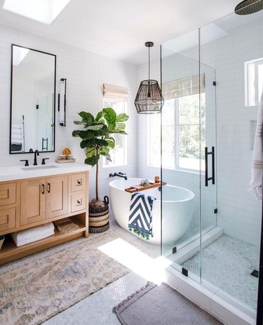 White bathroom with standalone bathtub, tall plant, wooden vanity, and black hardware