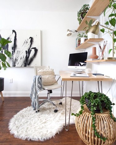 Minimal home office with blonde wood desk featuring metal hairpin legs, rolling beige swivel chair, floating wood shelves, and faux-fur white sheepskin rug
