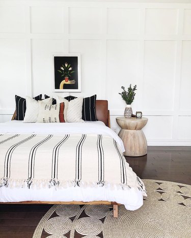 White bedroom with wood floor, wooden hourglass-shaped side table, bed with brown leather headboard and white bedding in addition to black-and-white striped throw pillows and blankets. 