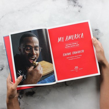 Hands holding an open book titled "My America: Recipes from a Young Black Chef: A Cookbook" with red pages.