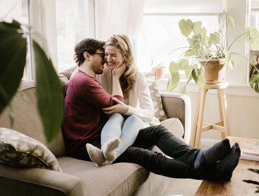 A white man and woman are sitting on a beige couch surrounded by sunny windows and plants. He has his feet resting on a coffee table in front of the couch, and she has her legs resting over his lap. He is touching her face, and she is smiling with her eyes closed.