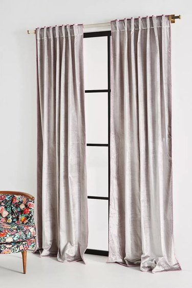 A pair of gray velvet blackout curtains in an all-white bedroom next to a colorful floral chair