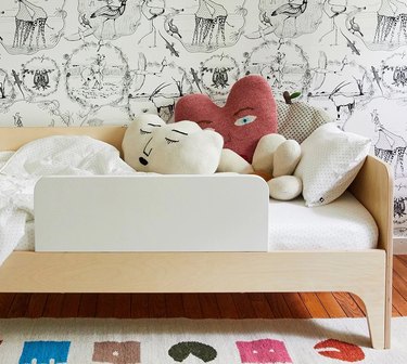 Wooden children's bed with white mattress and pillows with faces in front of a patterned wall.