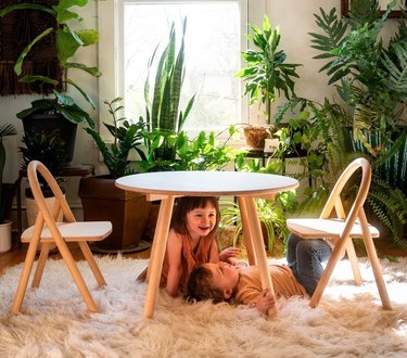A wooden table for children with two chairs in front of greenery with two toddlers laughing under the table.