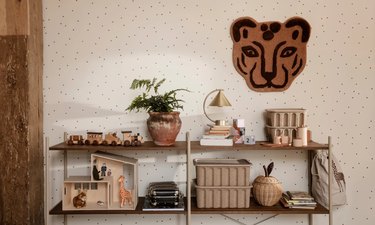 A muted shelf for children topped with books, toys, and plants in front of a beige wall with a tiger print.