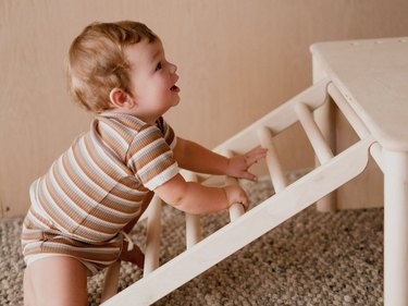 Baby in a striped jumpsuit climbing a wooden ladder that leads to a table.