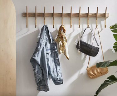 midcentury modern light wood coat rack with gold hooks against a white wall with coat, two purses, and keys hanging from it.