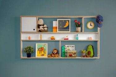 Mounted wall shelving unit full of children's books, stuffed animals, and toys on a dark green wall.