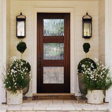 A farmhouse-style door with three glass horizontal panes is seen on a house with white siding; two planters with white flowers are on the porch as well as two matching black lanterns