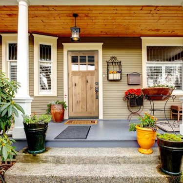 A light wood craftsman-style door on a large front porch that has many planters and a wooden ceiling