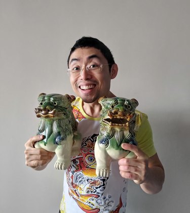Cliff tan holding two green lion sculptures in his hands while wearing an shirt featuring a yellow, orange, and white design.