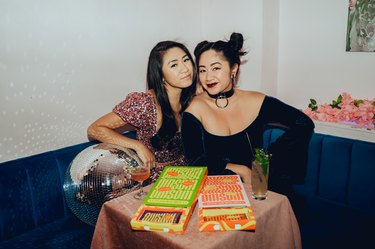 Vanessa and Kim Pham sitting on a blue velvet couch next to a disco ball behind a table featuring Omsom products.