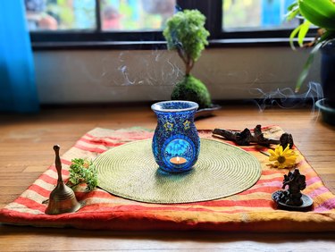A bell, an incense holder/lit incense, a diffuser/lit candle, a stick, a flower and plant cutting are on displayed on a colorful fabric on the floor