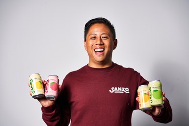 Sandro Roco in a dark red Sanza shirt holding four cans of Sanzo beverages in pink, yellow, and green.