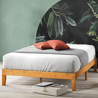 Zinus Alexia Wood Platform Bed Frame is in a room with green paint on the walls and gray wood floors. There are two red pillows and white and gray sheets on the bed.