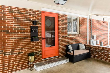 Brick house with an orange door, black sconce light, number plaque, black wicker bench, and gold plant stand